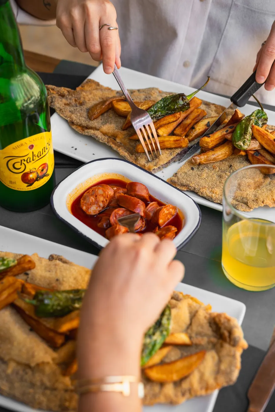 Comensal cortando un torto de maíz con patatas fritas y pimientos, sidra asturiana al fondo